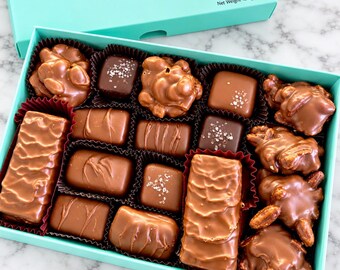May include: A turquoise box filled with an assortment of milk and dark chocolates, including nut clusters, sea salt squares, and rectangular bars. The box lid is labeled MALIBUGIFT Chocolaterie & Gifts with a net weight of 16 ounces.