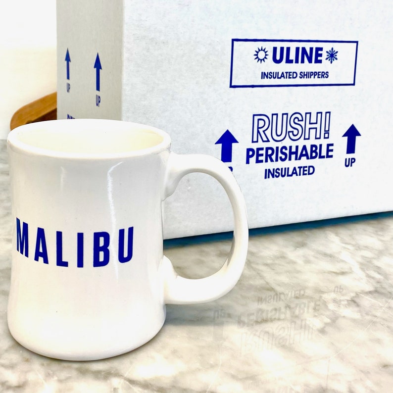 May include: White ceramic mug with a blue "MALIBU" logo. The mug is sitting on a marble surface next to a white box with blue text that reads "ULINE INSULATED SHIPPERS RUSH! PERISHABLE INSULATED UP".