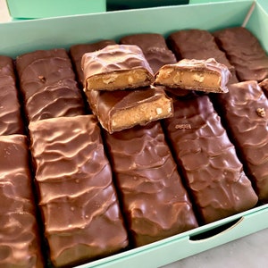 Op de afbeelding: Een doos met chocolade-karamelrepen, waarvan er een paar opengebroken zijn om de karamel en noten erin te laten zien. De doos is lichtblauw en de chocolade-repen zijn in rijen gerangschikt.