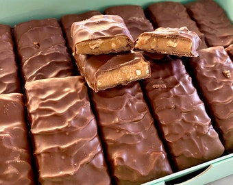 Peut inclure: Une boîte de barres chocolatées au caramel, dont certaines sont cassées pour révéler le caramel et les noix à l'intérieur. La boîte est bleu clair et les barres chocolatées sont disposées en rangées.