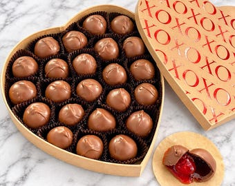 May include: A heart-shaped cardboard box filled with twenty-one round milk chocolate truffles in dark brown paper liners. The lid features a repeating red foil XOXO pattern. One truffle is cut open to reveal a bright red cherry and syrup center.
