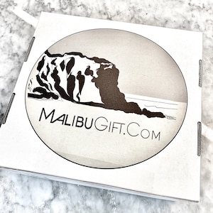 May include: A white cardboard box with a circular logo featuring a black silhouette of a cliff and ocean waves. The logo text reads "MALIBUGIFT.COM".