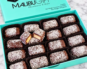 May include: A turquoise box labeled MALIBUGIFT Chocolaterie & Gifts containing rectangular chocolate candies topped with white coconut flakes. The box lid text states Net Weight 16 ounces (1 pound). One candy is broken to reveal a light tan interior.