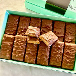 May include: A box of chocolate caramel candy bars. The bars are arranged in rows and are all wrapped in brown foil. The box is light blue with a white label that says "Eight 1 oz Net Wt."