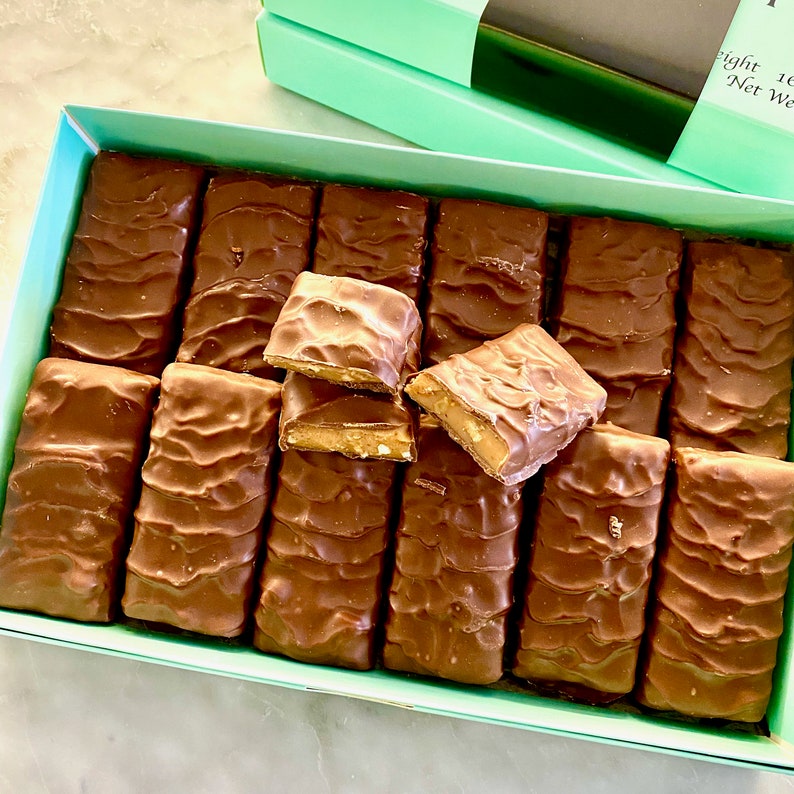 May include: A box of chocolate caramel candy bars. The bars are arranged in rows and are all wrapped in brown foil. The box is light blue with a white label that says "Eight 1 oz Net Wt."