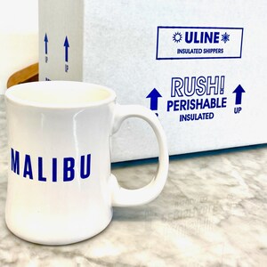 Op de afbeelding: Een witte keramische mok met een gebogen handvat en het woord MALIBU in blauwe hoofdletters op de voorzijde. Achter de mok staat een witte, ge&iuml;soleerde ULINE-verzenddoos met blauwe tekst die RUSH! PERISHABLE INSULATED en pijlen weergeeft.