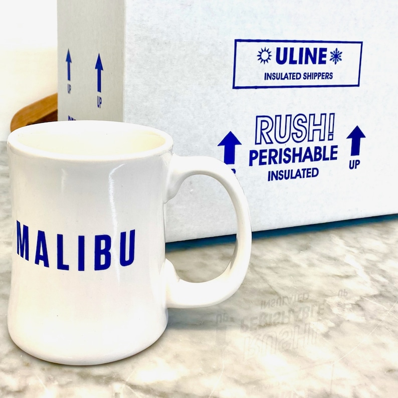Op de afbeelding: Een witte keramische mok met een gebogen handvat en het woord MALIBU in blauwe hoofdletters op de voorzijde. Achter de mok staat een witte, ge&iuml;soleerde ULINE-verzenddoos met blauwe tekst die RUSH! PERISHABLE INSULATED en pijlen weergeeft.