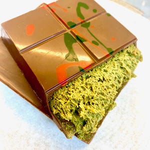 Op de afbeelding: Een chocoladereep met een groene vulling, mogelijk matcha. De donkere chocolade buitenkant is versierd met oranje en groene verfspatten. De reep is opengesneden en onthult de groene vulling. De reep ligt op een lichtgekleurd oppervlak.