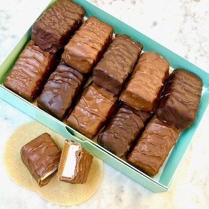 May include: A box of chocolate-covered candy bars with a light blue interior. The bars are various shades of brown, with some appearing to have a caramel or marshmallow filling. Two candy bars are cut open, revealing the white filling.