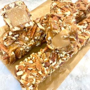 Op de afbeelding: Close-up van verschillende pecan praline repen. De repen zijn bedekt met gehakte pecannoten en hebben een karamelkleurige vulling. De repen liggen op een bruin papieroppervlak.