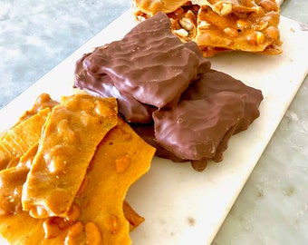 May include: A white marble serving board with three different types of peanut brittle. One type is a traditional peanut brittle, another is a chocolate peanut brittle, and the third is a peanut brittle with a caramel topping.