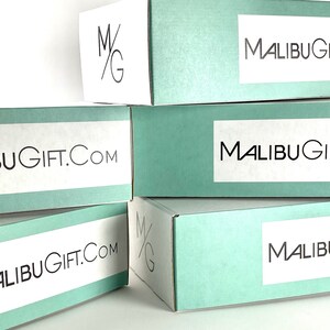 Op de afbeelding: Een stapel van vijf rechthoekige kartonnen verzenddozen in een lichtblauwe kleur. Elke doos is voorzien van een wit rechthoekig label met de zwarte tekst MALIBUGIFT.COM. Sommige dozen tonen ook een zwart M/G-logo op het zijpaneel.