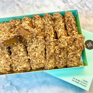 Op de afbeelding: Een lichtblauwe doos gevuld met rechthoekige Engelse toffeesticks bedekt met gehakte noten en chocolade. Het deksel heeft een zwart rond etiket met de tekst English Toffee Sticks en een nettogewicht van 454 gram.