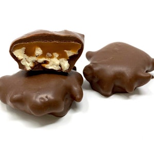 Op de afbeelding: Drie met chocolade omhulde snoepjes, waarvan er &eacute;&eacute;n doormidden is gesneden om een kern van goudkleurige karamel en lichtgekleurde stukjes noot te onthullen. De buitenkant is een gladde, stevige melkchocoladelaag met een onregelmatige vorm.