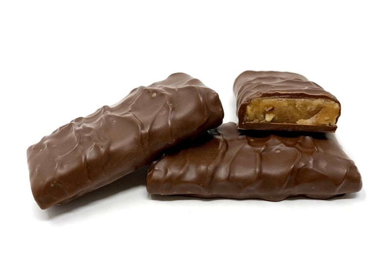 Op de afbeelding: Drie rechthoekige, met chocolade omhulde repen met een golvend oppervlak. E&eacute;n reep is doormidden gebroken, wat een lichtbruine, dichte en textuurrijke vulling onthult met kleine zichtbare stukjes noot in de melkchocolade omhulling.