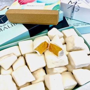 Op de afbeelding: Een doos gevuld met witte chocolade bedekte honingraat snoepjes. De snoepjes zijn in een enkele laag gerangschikt en sommige stukken zijn gebroken, waardoor de gele honingraat zichtbaar wordt.