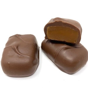 Op de afbeelding: Twee rechthoekige melkchocolades met een gladde, lichtbruine coating. E&eacute;n stuk is heel en vertoont subtiele inkepingen aan het oppervlak, terwijl de andere doormidden is gesneden om een stevige, dichte, lichtbruine karamelkleurige vulling te tonen.