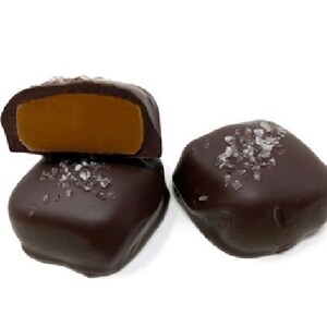 Op de afbeelding: Twee vierkante pure chocolade bonbons bestrooid met grove zeezoutkristallen. E&eacute;n bonbon is doormidden gesneden om een gladde, goudbruine karamelvulling te onthullen, terwijl de andere heel is en de pure chocoladelaag en het zout toont.