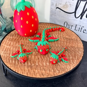 May include: A red and green strawberry-themed decorative set. The set includes a large strawberry-shaped object and three strawberry dragon figurines. The objects are arranged on a wooden tray with a black metal stand.