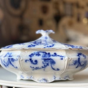 May include: A white porcelain tureen featuring a blue floral pattern with scrolling motifs. The oval-shaped serving dish includes two side handles, a matching lid with a decorative finial, and a scalloped base with gold-toned trim along the edges.
