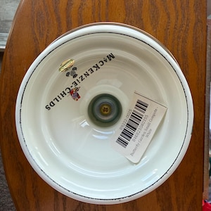 May include: A white compote bowl with a black rim, featuring the MacKenzie-Childs logo and a small butterfly graphic on the interior base. A barcode sticker with the text Butterfly Garden Small Compote - White is attached to the interior surface.