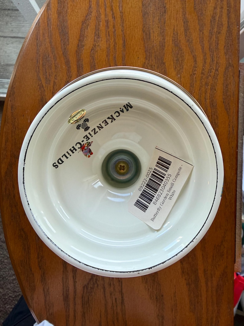 May include: A white compote bowl with a black rim, featuring the MacKenzie-Childs logo and a small butterfly graphic on the interior base. A barcode sticker with the text Butterfly Garden Small Compote - White is attached to the interior surface.