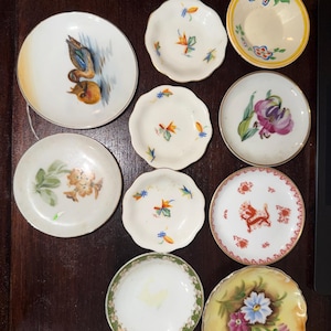 May include: A collection of small, decorative porcelain plates with various hand-painted designs. The plates feature floral, bird, and abstract patterns in a range of colors, including yellow, blue, pink, and green. The plates are arranged on a dark wooden surface.