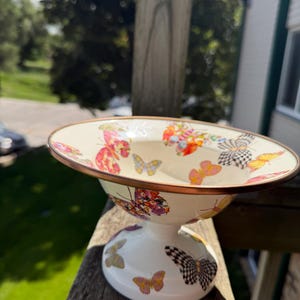 May include: A cream-colored ceramic pedestal bowl featuring a wide, flared rim with a metallic copper-toned edge. The exterior and interior surfaces are decorated with a repeating pattern of colorful butterflies in shades of pink, orange, yellow, and black.