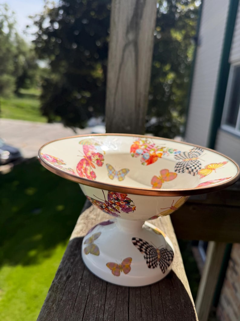 May include: A cream-colored ceramic pedestal bowl featuring a wide, flared rim with a metallic copper-toned edge. The exterior and interior surfaces are decorated with a repeating pattern of colorful butterflies in shades of pink, orange, yellow, and black.