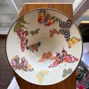 May include: A round, cream-colored ceramic bowl featuring a decorative pattern of various butterflies. The butterflies display diverse colors and wing patterns, including checkered, floral, and solid designs. The bowl has a thin, dark metallic rim.
