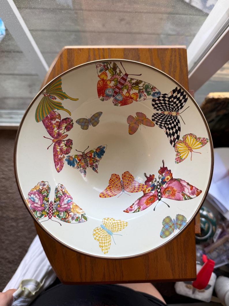 May include: A round, cream-colored ceramic bowl featuring a decorative pattern of various butterflies. The butterflies display diverse colors and wing patterns, including checkered, floral, and solid designs. The bowl has a thin, dark metallic rim.