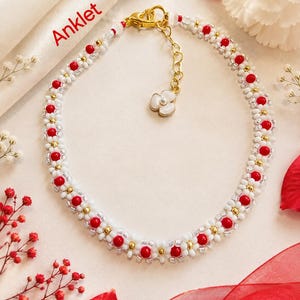 May include: An anklet featuring a repeating daisy pattern made of white and red beads with small gold-tone metallic accents. The piece includes a gold-tone lobster clasp, an adjustable chain, and a small white flower charm with a central bead.