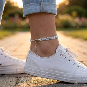 May include: An anklet featuring a repeating pattern of small white and light blue flower-shaped beads. The jewelry includes a silver-toned metal lobster clasp, an adjustable chain extension, and a small dangling heart-shaped charm at the closure.