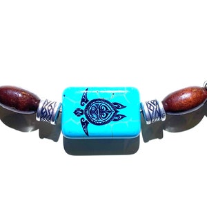 May include: A turquoise blue rectangular bead with a black turtle design is strung between brown wooden beads and blue and white porcelain beads. The beads are strung on a silver chain.
