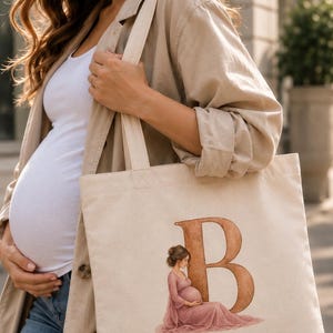 May include: A beige canvas tote bag featuring a large, brown, serif-style letter B. Inside the curve of the letter is a watercolor-style illustration of a pregnant person with brown hair wearing a long, flowing, dusty rose-colored dress.