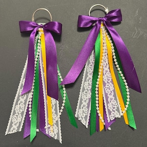 May include: Pair of Mardi Gras-themed earrings. Each earring features a purple satin bow, with green, yellow, and purple ribbons, white lace, and a string of white beads. The earrings have silver hoops.