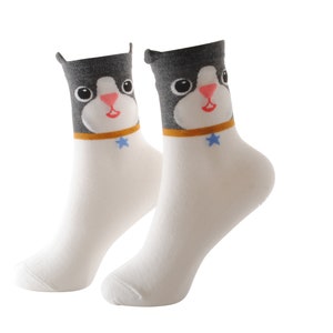 May include: A pair of white socks with a black and gray cat face design. The cat has a blue star on its collar.