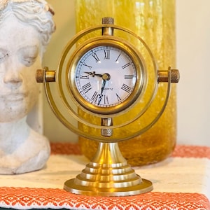 May include: A brass-toned metal desk clock featuring a circular frame that rotates on a central axis. The clock face is white with black Roman numerals and the word QUARTZ printed in black. It has a tiered circular base and two side knobs.