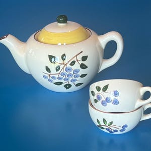 Könnte beinhalten: Eine weiße Keramik-Teekanne mit gelbem Deckel und dunkelgrünem Knauf, verziert mit einem Blaubeerzweig-Motiv. Zwei passende weiße Keramiktassen sind daneben gestapelt, jede mit einem ähnlichen Blaubeerzweig-Muster versehen.