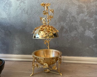 May include: A gold metal incense burner with a decorative floral design. The burner has a round bowl and a dome-shaped lid. The burner is on a wooden floor.