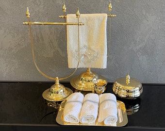 May include: A gold-toned towel rack with a cream-colored towel, a small gold cup, a gold tray with rolled white towels, and a gold container with a lid. The items are arranged on a black table.