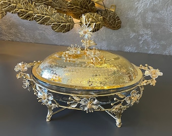 Può includere: Un piatto da portata dorato con un coperchio di vetro e accenti floreali. Il piatto ha tre gambe ed è decorato con dettagli intricati.