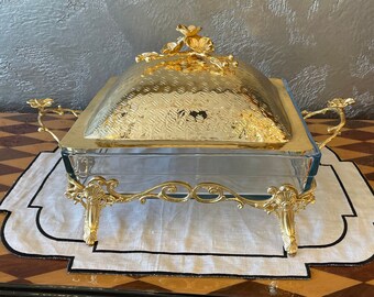 Può includere: Un piatto da portata dorato con base in vetro e coperchio decorativo. Il coperchio presenta un motivo floreale e la base ha gambe ornate.