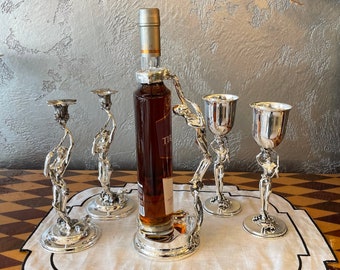 Puede incluir: Un juego de tapones de botella y copas decorativas plateadas con un diseño de figura. Los tapones de botella tienen forma de persona sosteniendo una botella de vino. Las copas tienen forma de persona sosteniendo una copa. El juego está sobre un mantel individual de lino blanco con borde negro.