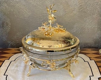 Può includere: Un piatto da portata placcato oro con una ciotola di vetro e dettagli floreali decorativi. Il piatto è su un sottopiatto di lino bianco e una superficie con motivo nero e oro.