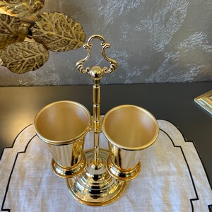 May include: Gold metal utensil holder with two cups. The holder has a decorative handle and a round base.