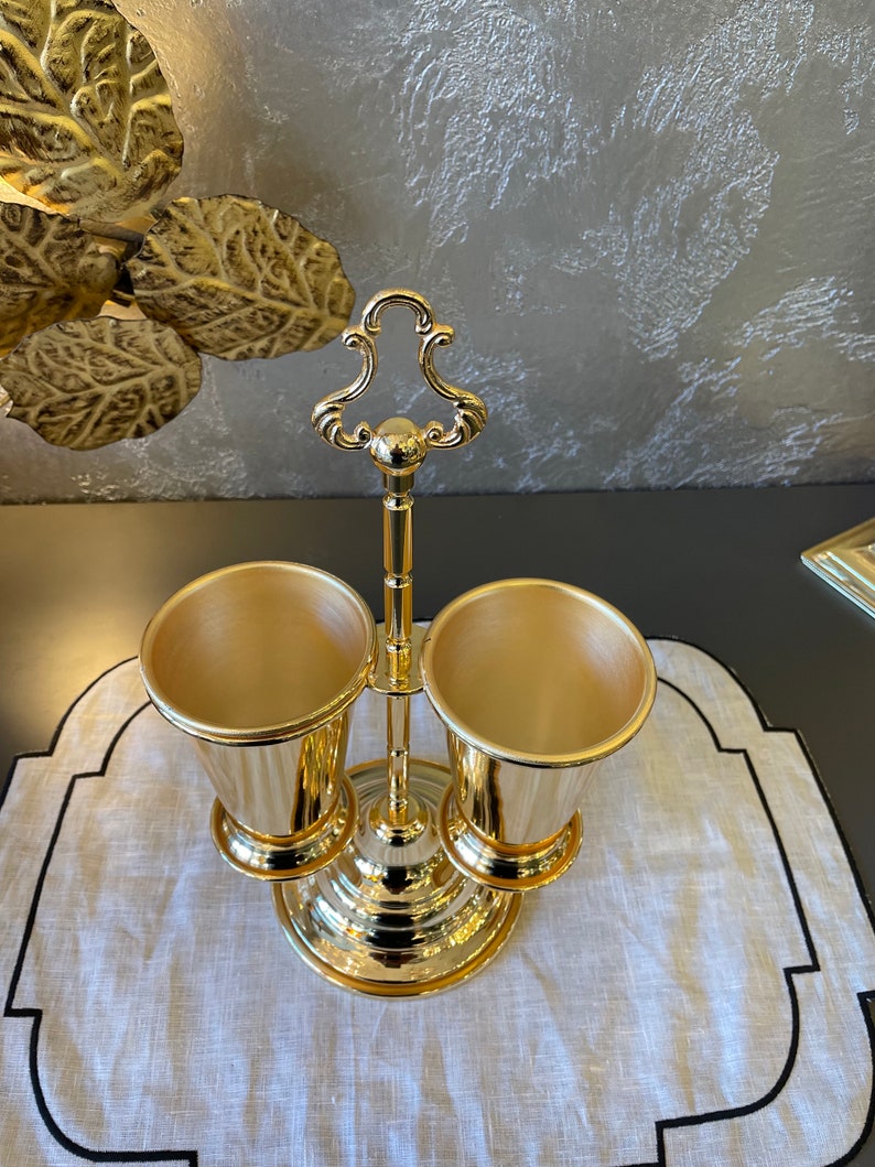 May include: Gold metal utensil holder with two cups. The holder has a decorative handle and a round base.