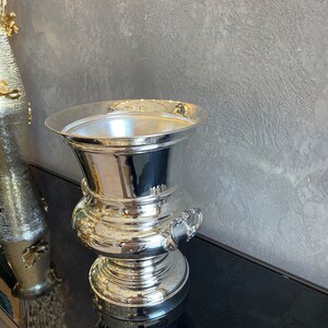 Puede incluir: Una cubitera de vino plateada pulida con una parte superior ancha y abierta y un cuerpo bulboso y escalonado. La cubitera tiene dos asas ornamentadas y refleja el entorno. Se encuentra sobre una superficie negra contra una pared gris texturizada.