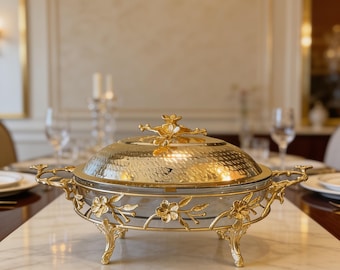 May include: An oval glass serving dish with a gold-toned metal frame featuring floral accents, branch-like handles, and four ornate legs. The dish includes a matching hammered gold-toned metal lid topped with a decorative floral finial.