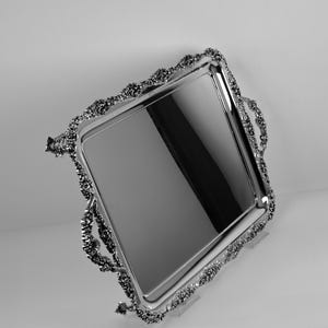 Puede incluir: Una bandeja rectangular plateada con bordes y asas ornamentados y texturizados. La bandeja tiene una superficie reflectante y el borde decorativo añade un toque de elegancia. La bandeja se utiliza probablemente para servir o exhibir.